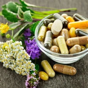 A bowl of various herbal supplement capsules surrounded by fresh medicinal herbs.
